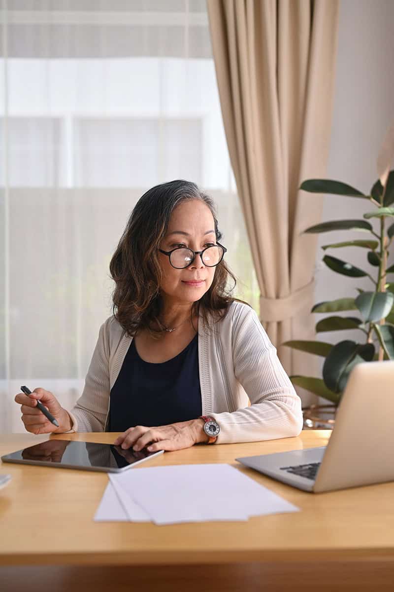 Middle-aged woman in eyeglasses working on a tablet at home.