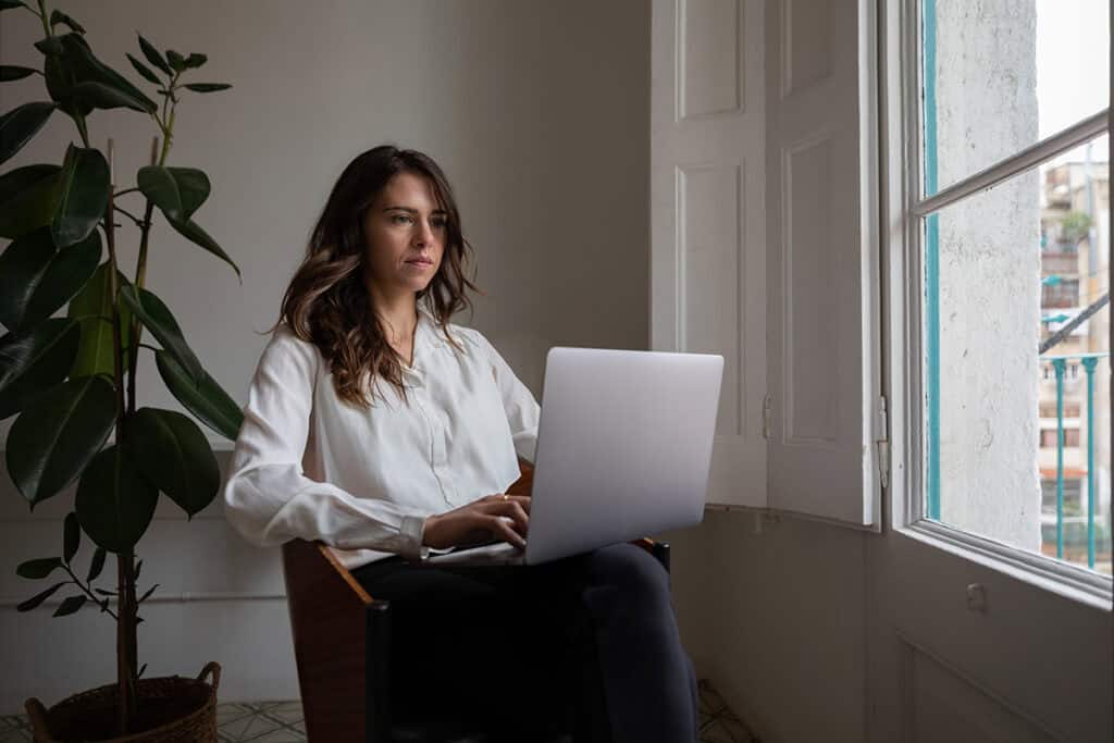 Professional woman working on her laptop at home, sitting near a large window with natural light.